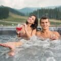A couple relaxing in an outdoor spa pool with drinks while overlooking scenic hills, ideal inspiration for first time spa pool buyers.