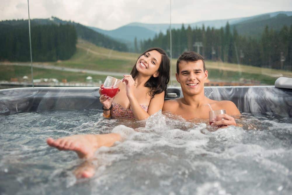 A couple relaxing in an outdoor spa pool with drinks while overlooking scenic hills, ideal inspiration for first time spa pool buyers.