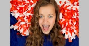 A cheerleader with long curly hair and braces holding red and white pom-poms, smiling energetically.
