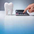 A close-up of a white tooth model next to a calculator, with a hand pressing a button. This visualises the financial cost calculation of dental procedures.